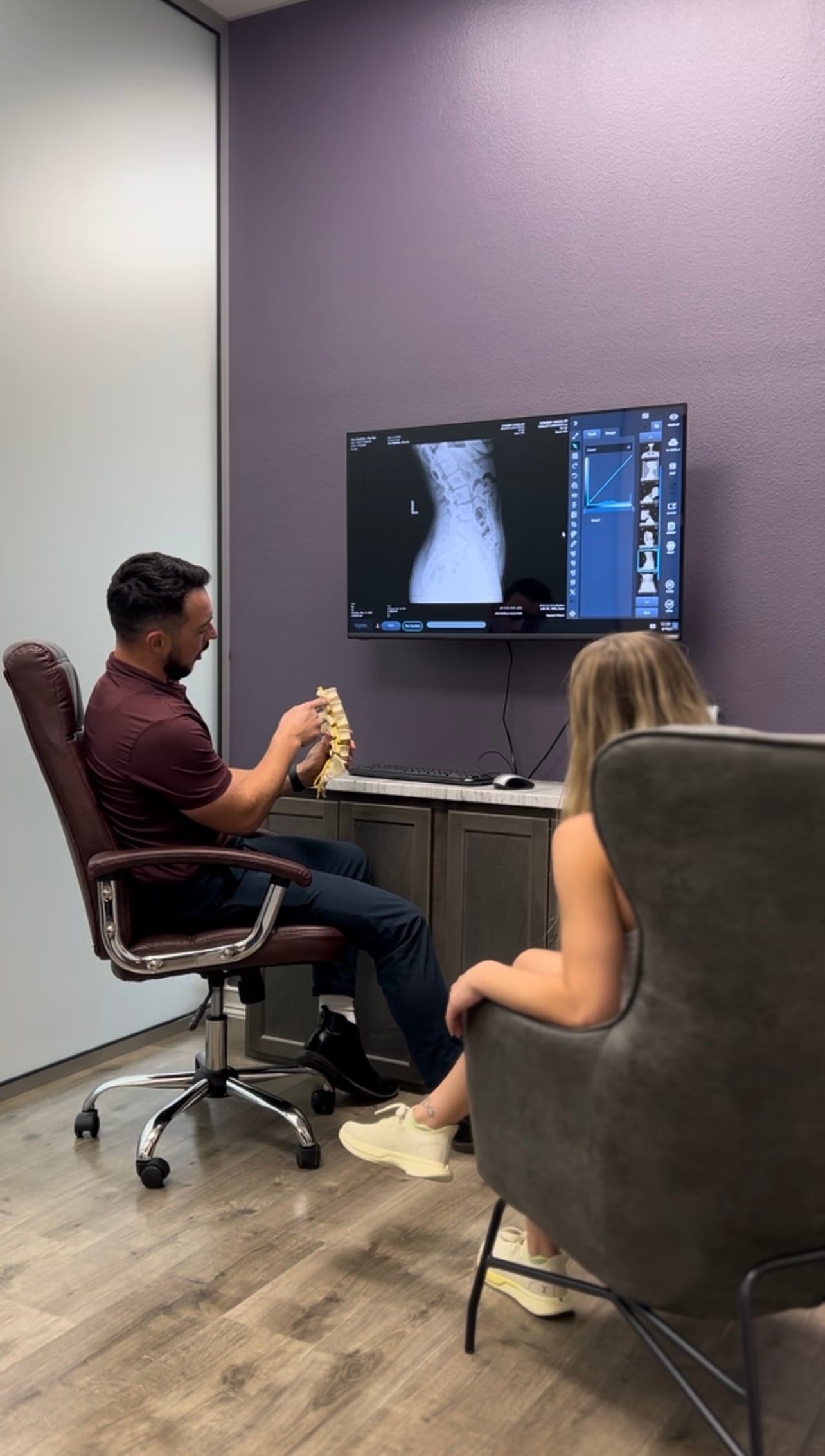 A male chiropractor points to a spine model while reviewing digital X-ray imaging during a consultation with a woman at Allied Wellness Centers in Waco, TX.